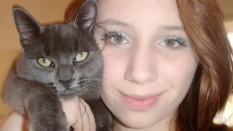 Family handout Alice looks straight at the camera and smiles. She has auburn hair. She is holding a grey cat up against her cheek