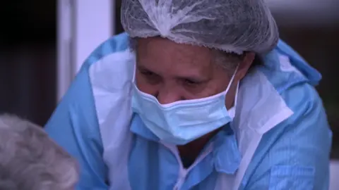 BBC A care worker in a face mask and other personal protective equipment leans towards a care home resident. This shot was taken in April 2020. 