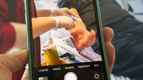 Getty Images A photograph of a phone filming a patient in hospital. In the viewfinder of the phone you can see a patient's arm and a nurse giving him a drug and performing a medical procedure. 