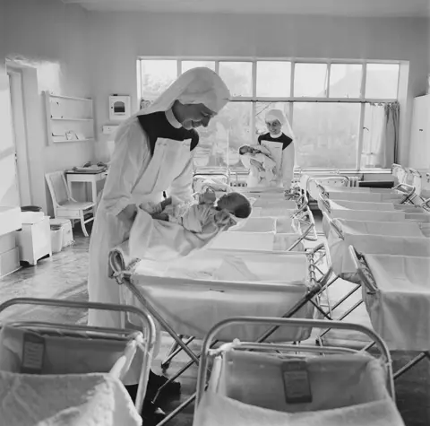 Hulton Archive/Getty Images A black and white photograph of cribs in a maternity ward. A nun wearing a veil is smiling and looking down at a baby she is holding. The cribs in front of the camera clearly display identity cards with the names and details of babies.