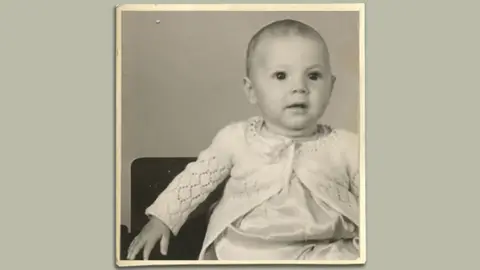 Family photo A black and white photo of Matthew's father as a baby in the 1940s. He is wearing a white knitted cardigan and staring off camera to the right.