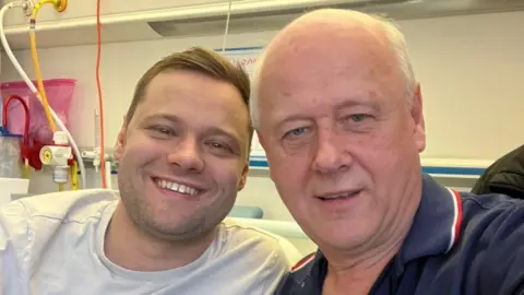 Brian Innes Kieran is smiling at the camera. He has short brown hair and is wearing a white t-shirt. Hi dad, Brian, is bald and is wearing a blue t-shirt.