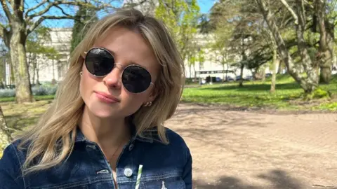 Eve Jones Eve Jones pictured in a dark denim jacket. She wears round sunglasses and looks at the camera. She has blonde mid length hair and wears small gold hoops. 