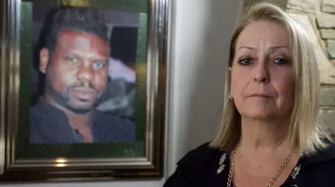 BBC Carole Charles, a middle-aged woman looking sad. A photo of her husband is visible on the wall behind her. She is made up smartly and wears a black top and a gold-coloured chain. She has blonde hair coming down to her shoulders.