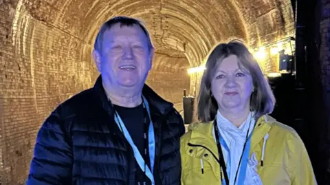 John Winnik John Winnik and Lyn Williams are standing in front of a lit tunnel. He is in a dark puffer jacket and she is in a yellow rain coat.