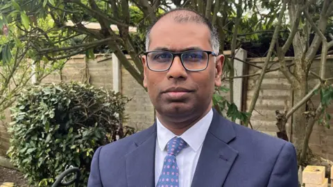 Profile shot of Dr Thanjavur Bragadeesh. He has short, dark, greying hair and black-rimmed glasses. He is pictured outside, with a fence and bushes behind him. He is wearing a dark blue suit, white shirt and blue tie patterned with pink circiles.  