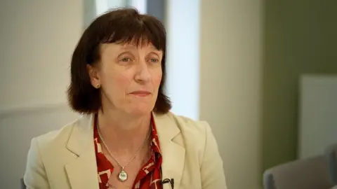 Durham County Council's director of public health Amanda Healy is sitting in a room with walls painted in muted pastels of while and pale green. The arm of a grey chair is out of focus behind her and to her left. She has chin-length dark hair with a fringe and is wearing a cream suit jacket over a red and cream shirt. She has a silver locket around her neck.