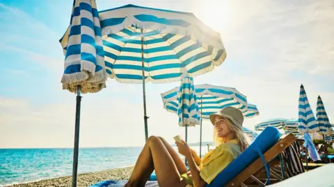 Getty Images A woman sits on a sun lounger under a sun umbrella on a beach. 