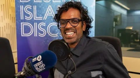 A picture of comedian Romesh Ranganathan with a microphone that has Desert Island Discs branding on it.