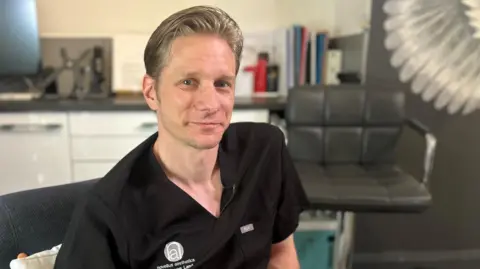 Dr Steven Land. A man in his 40s with blonde hair wearing a black, medical scrubs-style top with his company's logo on one side. He is sitting on a grey arm chair in an aesthetic practitioner's room. Drawers and another chair are out of focus behind him.