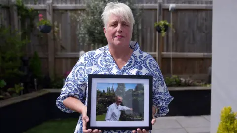 Tracy Fisher, holding a black-framed framed picture of her father, Brian. She has short grey hair and is wearing a blue and white patterned dress. She is pictured on a garden patio, with fences, raised beds and hanging baskets behind her. In the photograph she is holding, her father is smiling at the camera while pointing at something in the garden to the side of him.