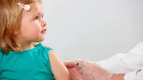 Getty Images A young child wearing a green top has her sleeve rolled up and is ready to get a vaccine from a doctor (unpictured) whose arms and hands are in view, who is holding a vaccine