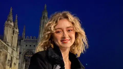Gabriel & Sebastian Shemirani Paloma, smiling at the camera as she sits on the wall outside King's College, Cambridge, with the windows of the chapel illuminated and a dark blue twilight sky behind her. She is wearing a warm black jacket but has bold make-up with pink eyeshadow for a night out.