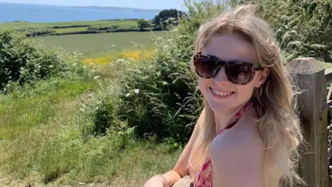 Eve Jones Picture of Eve Jones, as she stands by a wooden gate overlooking rolling green hills in front of the sea. She wears a pink halterneck top and looks over her shoulder smiling with her teeth. She has light blonde mid length hair, she wears square tortoise shell sunglasses with silver small hoops. 