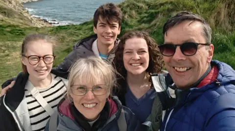 Paul Silvester Paul and his family - wife, two daughters and son - all smiling while they are out for a hike with cliffs and the sea in the background 