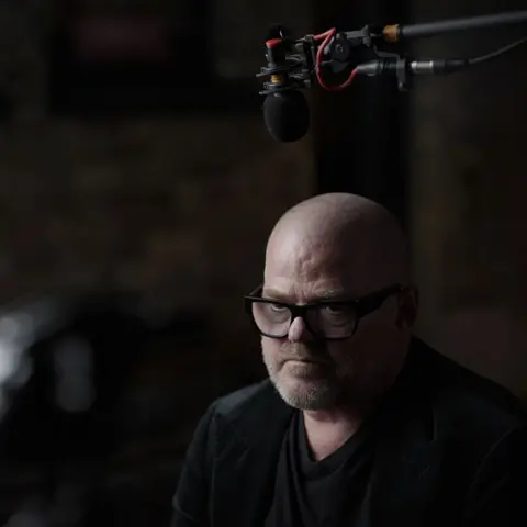 Heston Blumenthal pictured during an interview, he is wearing black frame glasses a black t-shirt and jacket and there is a microphone above his head.