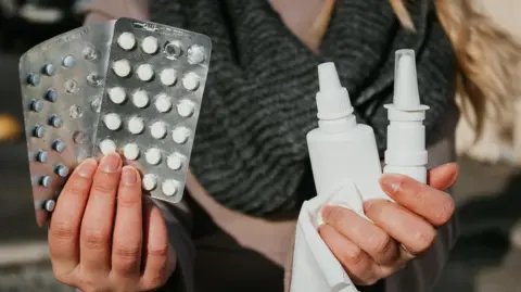Getty Images A woman holds up two packs of antihistamine pills in one hand and nasal and eye sprays on in the other, along with a tissue