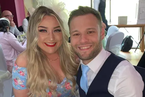 Kieran Innes Lara is wearing a light blue dress with flowers on it. She has long blonde hair and is wearing a fascinator in her hair. She is smiling at the camera and has red lipstick on. Kieran is in a waistcoat and light blue tie. He is smiling at the camera. They are at a wedding.