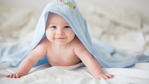 Getty Images A smiling baby lies on a bed wearing a light blue hooded towel.