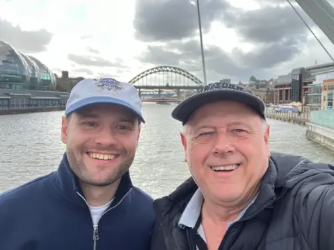 Brian Innes Kieran is smiling at the camera. He is wearing a dark blue fleece jacket and cap. His dad, Brian, is also wearing a cap and puffa jacket. The Newcastle bridge and Tyne River are in the background.