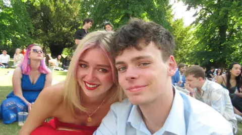 Ander Harris Paloma Shemirani, with a streak of pink in her hair and red lipstick, wearing a red off the shoulder outfit, leaning in close to Ander Harris, a white man with brown hair, tousled on top, wearing a light coloured collared shirt. They are both smiling as they sit on the grass, surrounded by trees and other young people, presumably students.
