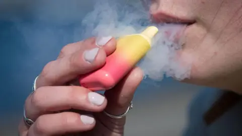 Getty Images A woman holds a yellow and red coloured vape to her mouth with a cloud of vapour surrounding it.