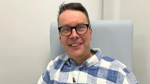 BBC Paul Silvester smiling in a chair wearing a blue and white checked linen shirt and black thick-rimmed glasses