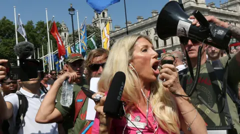 Getty Images Kate Shemirani, who has long blond hair and wears a pink outfit with what appears to be a stethoscope around her neck, at an anti-lockdown protest in 2021. A man nearby holds up a megaphone speaker as she speaks into the microphone part, which she holds in her hand. Behind her are flags on what looks like Parliament Square, along with crowds of people, some holding up cameras which they are pointing at the press photographer.