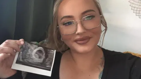 Natasha Major Natasha is smiling at the camera, wearing a black top and glasses, with long blonde hair, and holding a print-out of her pregnancy scan