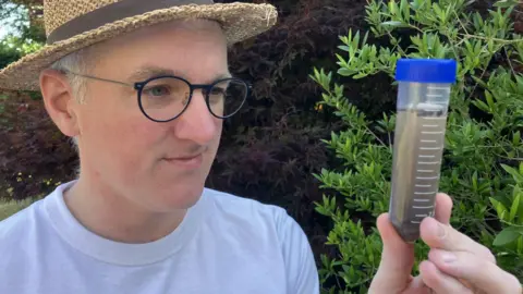 BBC James Gallagher wearing a white T-shirt and summer hat, looking closely at a vial containing pond-water