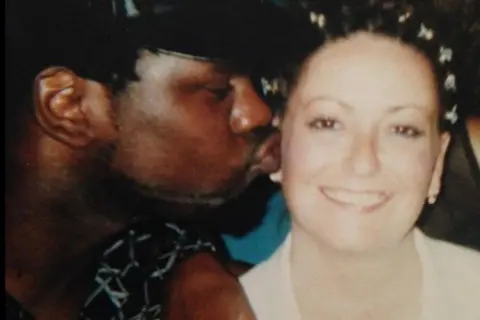 Charles family Winbourne and Carole Charles, seemingly at a party or event. Mr Charles is kissing the side of Mrs Charles's face as she smiles at the camera. She has decoration in her hair, pearl earrings and a white top. He is wearing a black shirt with a white pattern on it.