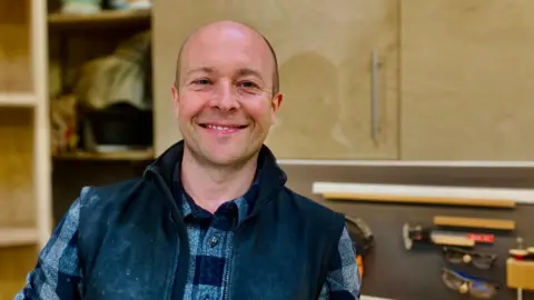 BBC Furniture maker Nick James is in his furniture making workshop in Newcastle.  He is looking at the camera and smiling and wearing his casual clothing. Behind him you can see his wood-working tools on the wall.           