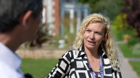 Vicky Bartlett, who has long blonde wavy hair and is wearing a black and white patterned top and purple lanyard.