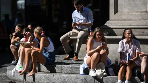 Getty Images Men and women sit on the steps of a fountain in the sunshine while eating their lunch and checking their phones. A number of women are wearing shorts. 