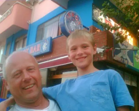 Brian Innes Brian Innes has his young son in his arms while on holiday. They are tanned and smiling and both have light blue tops on.