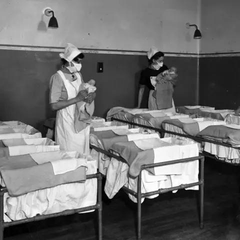 Hulton Archive/Getty Images A black and white photo of two nurses holding babies in a maternity ward in 1947. In the foreground are sixteen different cots with iron bed frames. It is impossible to see from the photo if they all contain babies.