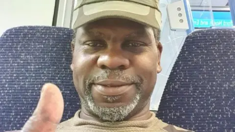 Charles family Winbourne Charles, a middle-aged man wearing a khaki cap and a beige top. He is putting one thumb up and appears to be sitting in a train carriage.