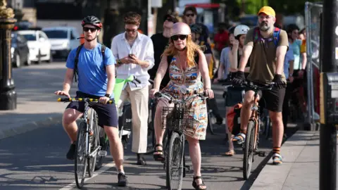 PA Media Cyclists pause on the road, some wearing helmets, as the sun shines down.