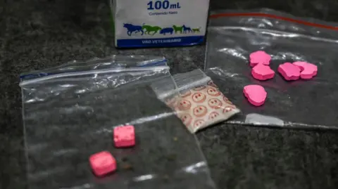 Getty Images A stock image of some illegal drugs including two bags with pink tablets inside and a bag of white powder in a plastic baggy with smiley faces on