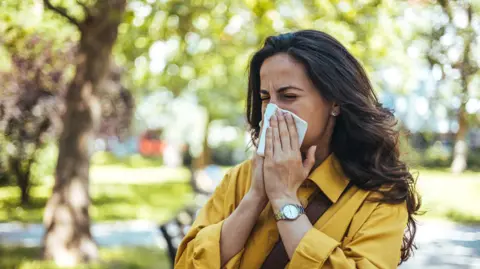 Getty Images A woman with long brown hair, wearing a yellow shirt, holds a tissue to her nose as she sneezes, with trees in the background