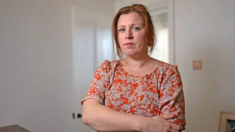 BBC Profile picture of Tassie Weaver. She has straight red hair which is tied back with some tendrils around her face. She is wearing a short-sleeved orange top, patterned with grey flowers and has her arms folded. She is pictured indoors, with a white open door behind her. 