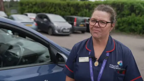 Angelina Blair, who is wearing a dark blue nurses uniform, has brown hair tied back and is wearing brown-rimmed glasses.