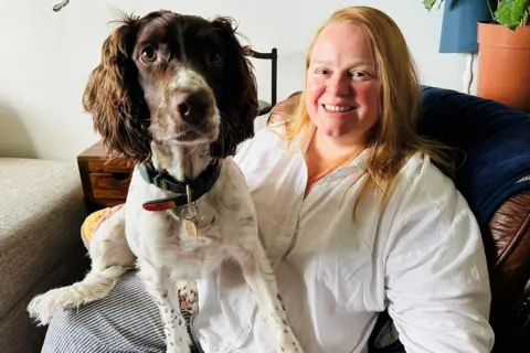 Woman, Marlene Lowe, with blonde hair, and white blouse, smiling at camera, with brown and white cocker spaniel dog on her lap.