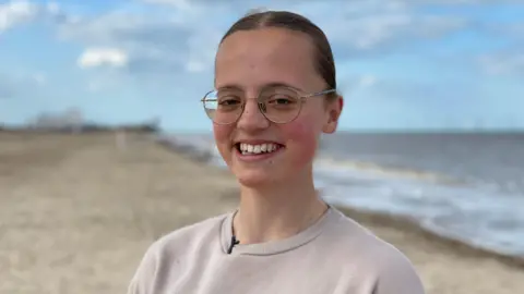 BBC Mary Catchpole, smiling with her hair tied back and wearing glasses. She is standing on a beach near her home in Norfolk