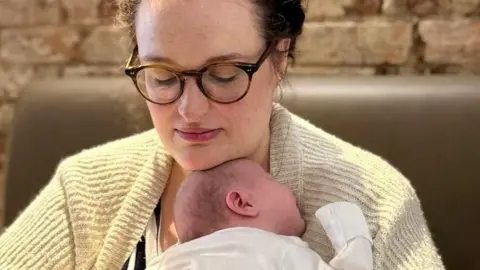 Charlie Beattie Charlie holds her baby June on her chest, while wearing a yellow cardigan. The baby is wearing a white baby suit