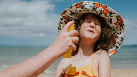 Getty Images A little girl on the beach wearing a sun hat and orange swimming costume grimaces as sunscreen is applied to her face. 