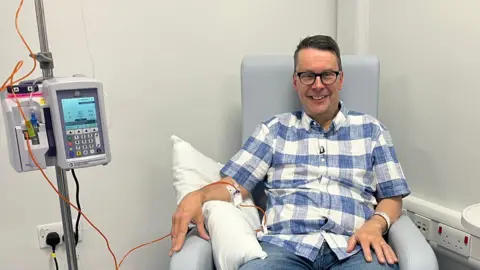 Paul wearing a blue and white checked shirt is sat in a chair, bare arm is resting on a cushion while an orange tube connects a cannula to a pump. 