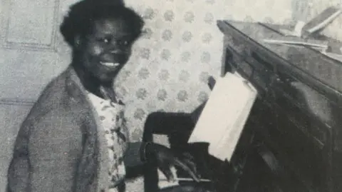 Other Grace Adebowale is pictured in her 40s in a black and white photo. She is smiling at the camera while seated at a piano which she appears to be playing.