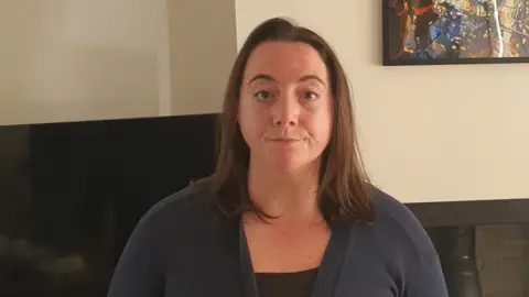 Beth Rodgers Beth Rodgers, a 32-year-old woman. She is standing in her living room wearing a navy cardigan and black top.