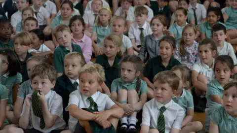 BBC Primary school pupils sit and listen, some with quizzical expressions.